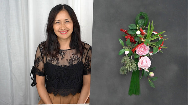 Instructor beside a New Year hanging arrangement featuring pink clay camellias, green bamboo leaves, red accents, and a long green tassel.