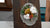 Christmas wall decoration with orange slices, white cotton flowers, red berries, pinecones, and green leaves on a gray plaque and dark wood backing.