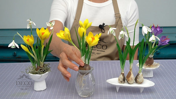 Instructor displaying spring clay flower arrangements with yellow and purple crocuses, white snowdrops, green leaves, and exposed bulbs.