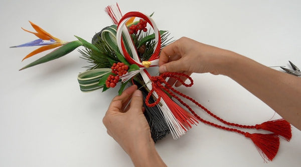 Hands assembling a Japanese New Year wall decoration with a clay bird-of-paradise flower, red berries, green leaves, and red-and-white cords.