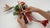 Hands assembling a Japanese New Year wall decoration with a clay bird-of-paradise flower, red berries, green leaves, and red-and-white cords.