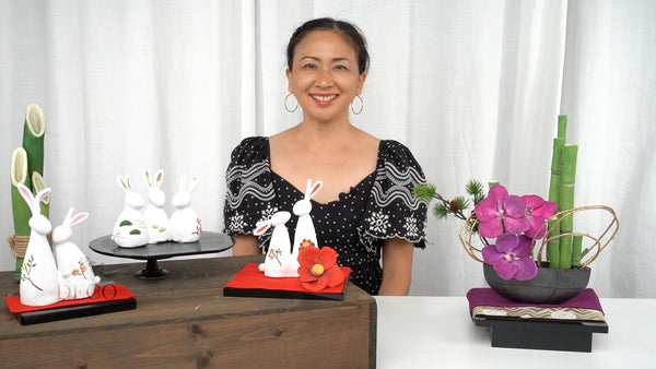Instructor displaying white clay zodiac rabbits and a New Year arrangement with purple Vanda orchids and green bamboo.
