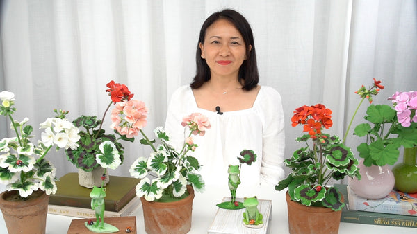 Instructor displaying potted clay geraniums with red, pink, and white flowers, green frog figurines, and small ladybugs.