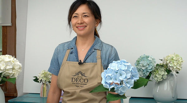 Instructor holding a blue clay hydrangea stem beside vases of white, pale green, and blue hydrangeas.