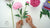 Hands holding a long-stemmed pink clay peony, with additional peony blooms, a bud, and green leaves on a white work surface.