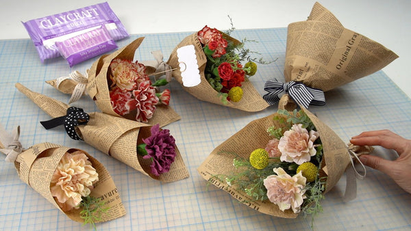Clay carnation bouquets in cream, pink, red, and purple, wrapped in printed brown paper with ribbons, greenery, and yellow Billy Buttons.⠋