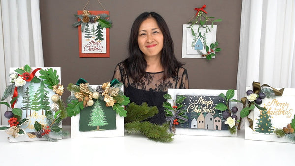 Instructor behind framed Christmas decorations embellished with clay flowers, holly, berries, acorns, foliage, and festive ribbons.