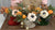 Christmas clay arrangements in red, cream, and green knitted-style boots, with white anemones, orange slices, berries, foliage, and gold accents.