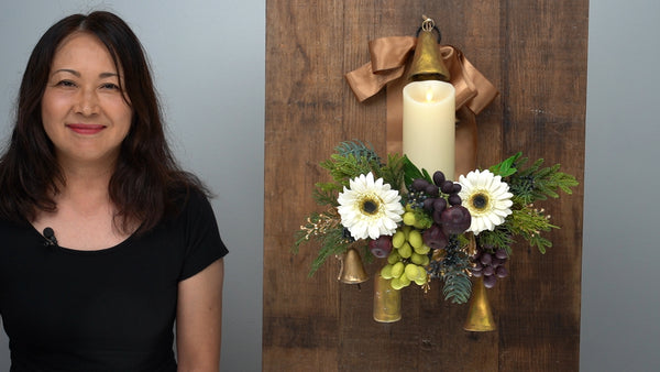 Instructor beside a Christmas wall arrangement with a candle, white clay gerbera daisies, green and purple grapes, figs, gold bells, and evergreen foliage.