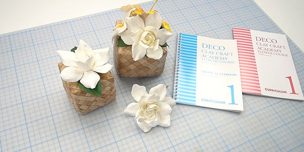 White clay gardenia flowers in woven containers beside DECO Flower Course 1 and Tropical Course 1 curriculum books.