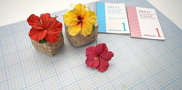 Red and yellow clay hibiscus flowers in woven containers beside DECO Flower Course 1 and Tropical Course 1 books.
