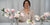 Instructor displaying handcrafted pink and white clay magnolia blossoms on leafy branches arranged in clear glass vases.