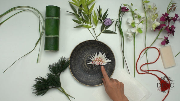 New Year clay project elements with a green bamboo-style vase, white and purple orchids, Fritillaria flowers, a crane ornament, pine foliage, and red cord.