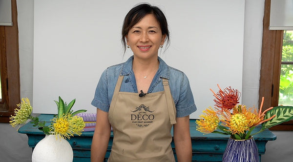 Instructor standing between vases of yellow, orange, and red clay pincushion proteas with green leaves.