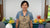 Instructor standing between vases of yellow, orange, and red clay pincushion proteas with green leaves.