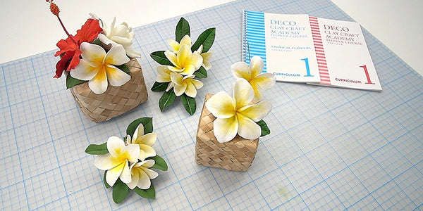 Three small woven baskets decorated with artificial plumeria flowers and green leaves, arranged on a light blue grid mat next to a blue and pink 'Decor' booklet.
