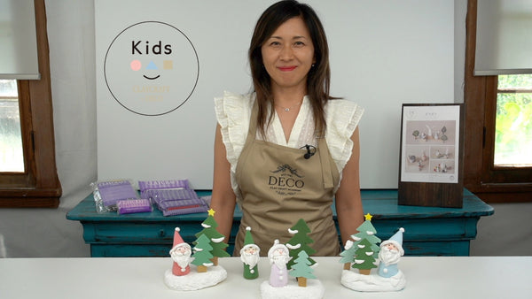 Instructor behind clay Santa figures in colorful outfits and snow-tipped green Christmas trees on white bases.