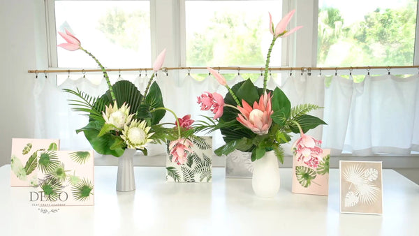 Tropical clay flower arrangements with pink and cream King Protea, Medinilla, banana flowers, and green foliage beside leaf-stenciled framed canvases.