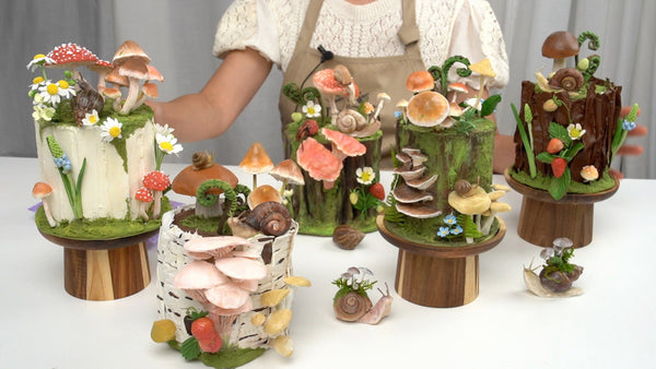 Woodland-themed clay cake sculptures decorated with mushrooms, daisies, strawberries, ferns, and snails.