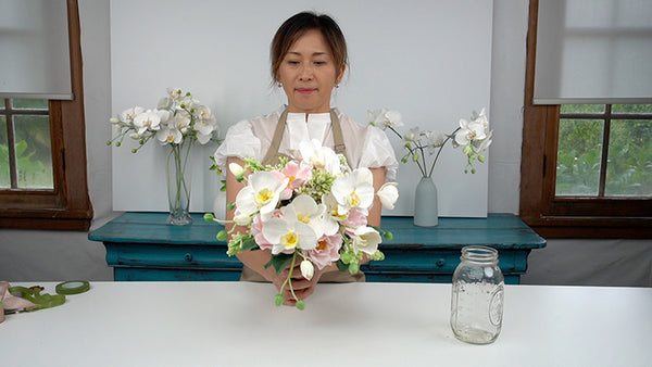 Instructor standing between a clay wedding bouquet of white orchids and pink flowers and white phalaenopsis orchid stems in a pale blue vase.