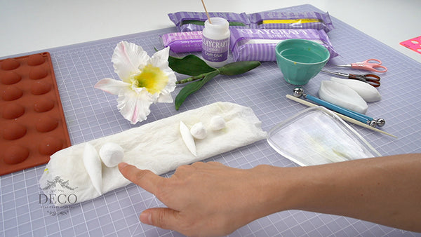 White clay cattleya orchid with a yellow center beside silicone molds, clay, and flower-making tools on a gridded work mat.